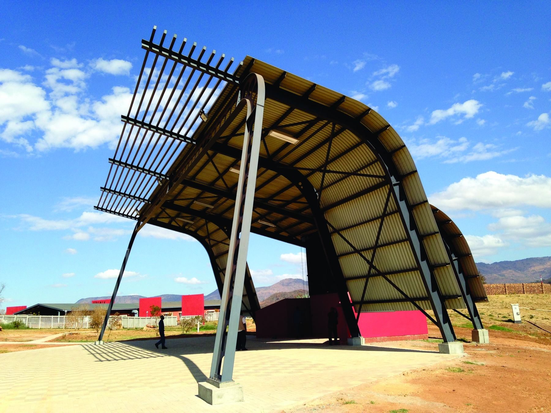 Curved steel canopy with a louvred screen on a tall frame, in a paved public space.