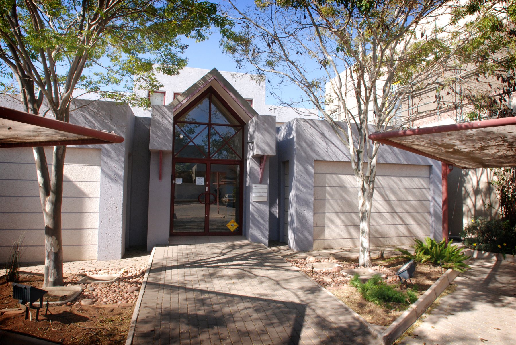 Entrance with a pitched, glazed gable and red-painted steel frame, set between textured plastered walls and trees.
